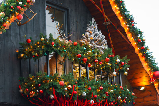 Festive Christmas wooden chalet balcony decorated with shining lights, illuminated evergreen garlands, ornaments, ribbons and a small tree, cozy winter holiday atmosphere with rustic architecture.