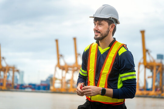 Port worker man wearing safety gear at shipping terminal, male engineer wearing safety vest and hard hat holding walkie talkie at industrial container port and shipping yard