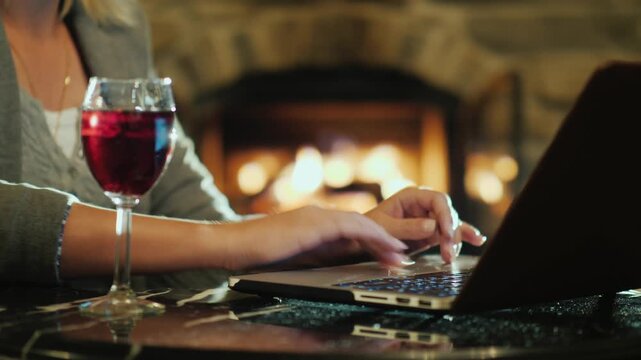 Business woman working on laptop at night with red wine glass by burning fireplace in cozy home interior.