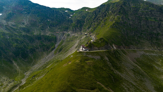 Aerial view of the winding Transfagarasan road cutting through the lush green Carpathian Mountains with a mountain lodge perched on the ridge in Curtea de Arges, Judetul Arges, Romania.