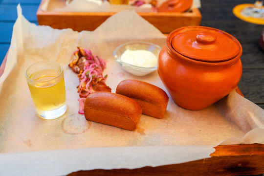 Hearty homemade soup served rustic clay pot, accompanied by small fresh bread rolls, crispy bacon pieces, and a cup of sour cream.