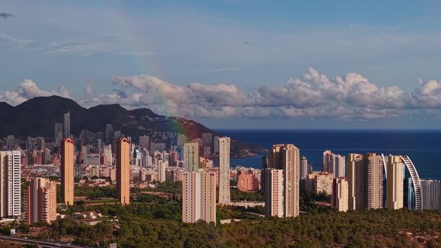 Cinematic aerial view of Benidorm skyline featuring colorful rainbow over high rise buildings and mountains.