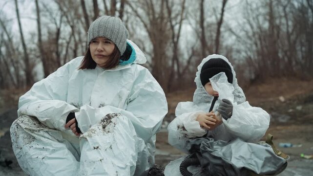 Mother child protective suits sharing food on cold roadside, bundled in knitted hats and gloves, child sipping from cup while adult offers comfort, muddy littered landscape with bare trees, intimate