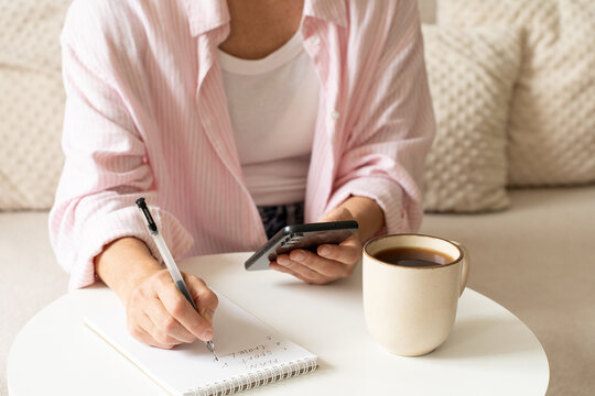 Woman enjoys leisurely home ritual, making notes in notebook while holding her smartphone and sipping coffee. A mindful approach to life, journaling, me-time, and balanced daily routine