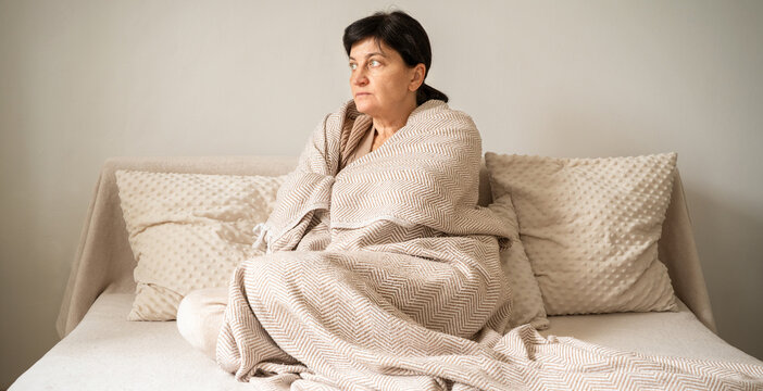 Mature woman sits on beige sofa, wrapped tightly in beige fringed throw, looking away with pensive and slightly anxious expression. Women's mental and hormonal health