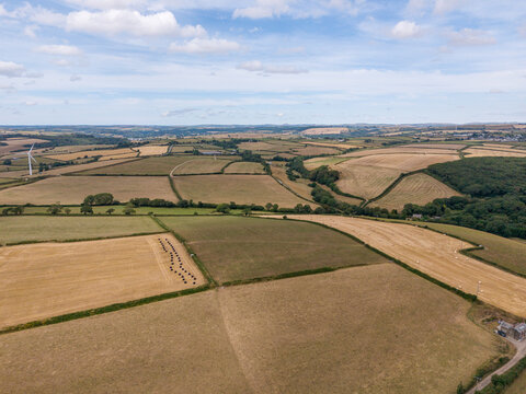 Aerial view of agricultural farmland featuring harvested fields, hedgerows, tracks, and a wind turbine under a blue sky with clouds in Looe, Cornwall, United Kingdom.