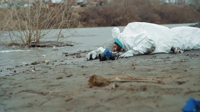 Child scientist collapsed on muddy shore wearing white protective suit, frost coating fabric, river water lapping nearby, scattered debris and plastic, abandoned industrial skyline in background,