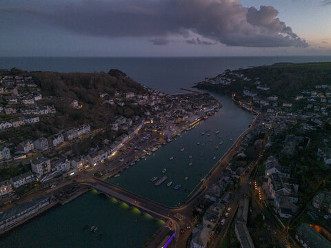 Aerial view of the coastal town of Looe at dusk, featuring the Banjo Pier, moored boats in the river estuary, and surrounding hillsides with residential buildings in Looe, Cornwall, United Kingdom.