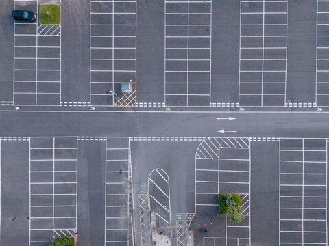 Aerial view of an empty asphalt parking lot with white painted parking spaces, directional arrows, and small green patches in Looe, Cornwall, United Kingdom.