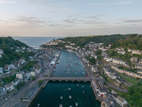 Aerial view of the coastal town of Looe, featuring the river estuary, small boats moored in the harbor, historic buildings, and lush green hills in Looe, Cornwall, United Kingdom.