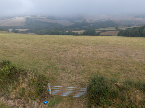 Aerial view of the misty rural countryside in Looe, featuring rolling agricultural farmland, foggy fields, and a metal farm gate in the foreground, Looe, Cornwall, United Kingdom.