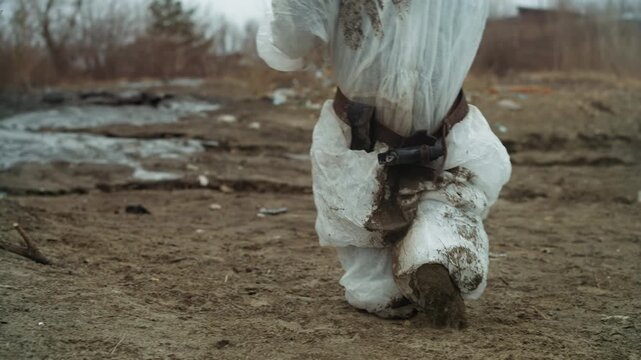 Person trudging in muddy protective suit, slow heavy steps across contaminated wasteland with caked mud on boots and lower suit closeup of stained gait conveys exhaustion, tension, and urgent escape