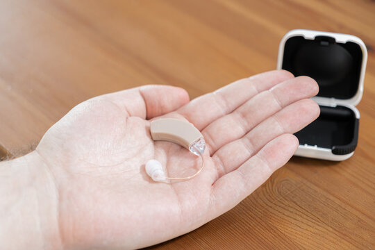 A modern beige digital hearing aid is held in a human hand near its storage case on a wooden table to illustrate healthcare, auditory assistance, and medical tech for the deaf