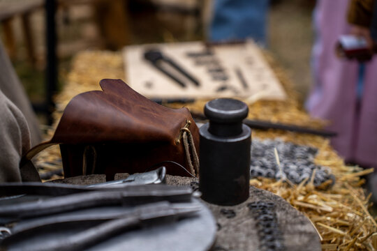 Authentic leather pouch, miniature anvil, and blacksmith pliers placed on wooden stump and straw bale at historical reenactment camp.