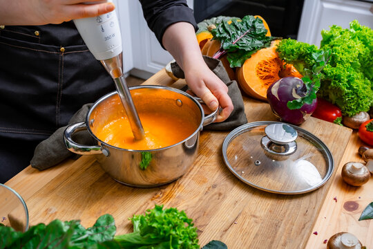 Comfort autumn food &mdash; chef hands blending hot pumpkin soup in metal pot. High haute cuisine meal preparation using modern kitchen appliance. Healthy eating lifestyle with seasonal organic dish.