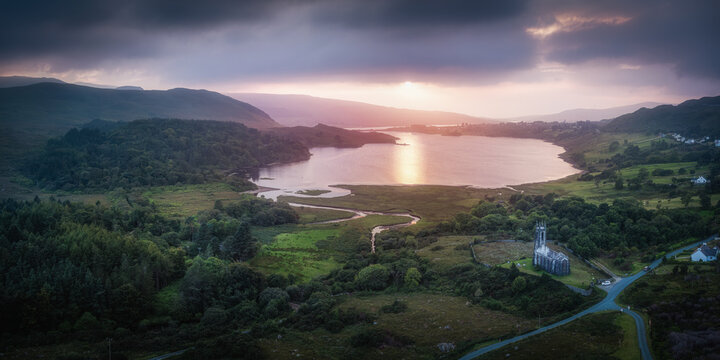 Aerial view of the historic Dunlewy Church ruins by the tranquil waters of Dunlewy Lough at sunset, surrounded by lush green hills in Dunlewy, County Donegal, Ireland.