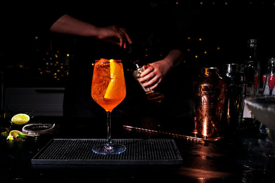 Orange aperol spritz cocktail with ice and fruit slice on dark bar counter - alcoholic drink mixology. Bartender preparing refreshing summer beverage in background.