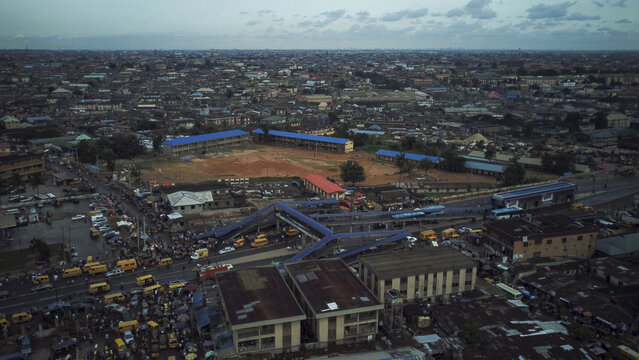 Aerial view of Agege featuring a pedestrian bridge over a busy road with yellow buses, school buildings with blue roofs, and dense urban sprawl in Lagos, Lagos, Nigeria.