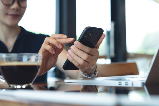 Young asian woman using smartphone with laptop on table during remote work at coffee shop, digital nomad lifestyle, online business concept
