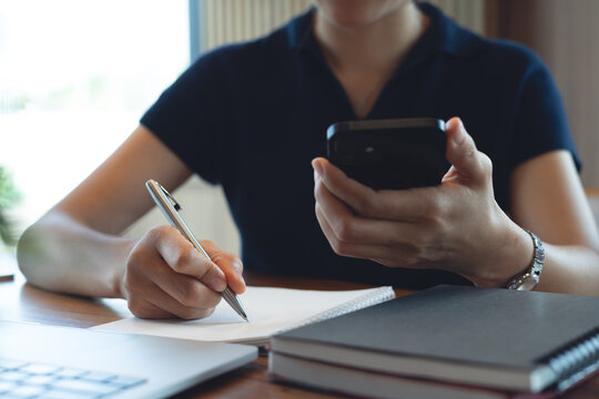 Woman using smartphone searching the information and writing in notebook with laptop on table, remote working at coffee shop, online business concept
