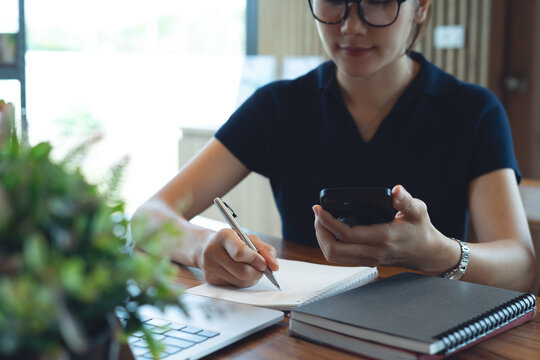 Asian business woman using smartphone searching the information, writing in notebook with laptop on table, remote working at coffee shop, online business concept