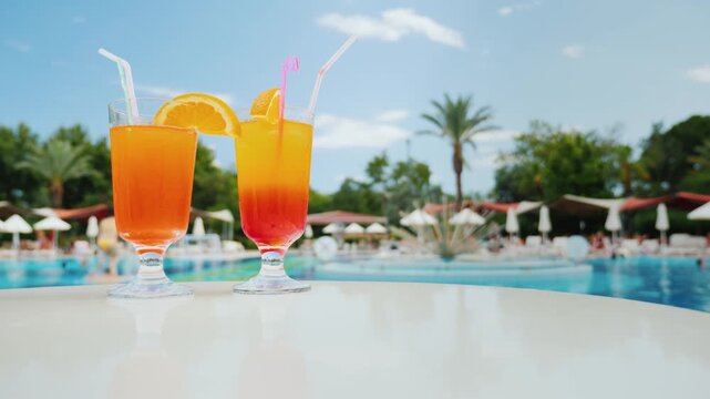 Two colorful cocktails with orange slices and straws on table near pool with people swimming.