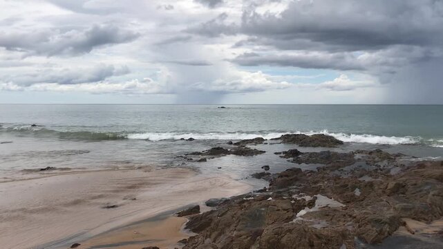 Smooth panning shot of tropical beach with ocean waves ,rocky shoreline and dramatic cloudy sky.Natural coastal scenery with moving sea wave and an overcast atmosphere in Thailand