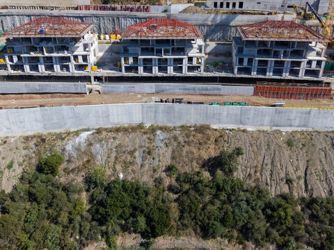 Aerial view of a construction site featuring three modern residential buildings with red roof frames, a large concrete retaining wall, and a hillside with trees in Bursa, Bursa, Turkiye.