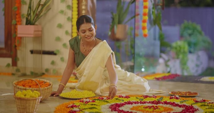 South Indian young adult woman lady wear saree sitting on floor make floral good rangoli indoor home place. Happy female girl arrange flower petals design enjoy diwali festival at night house