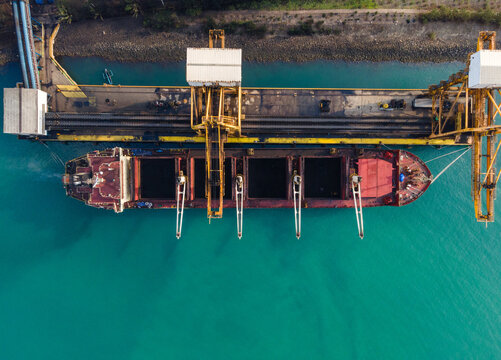 Aerial view of a large cargo ship docked at a port facility with industrial loading cranes and turquoise water in Cilacap, Central Java, Indonesia.