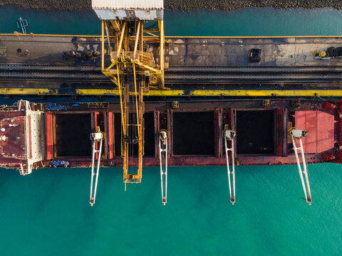 Aerial view of a cargo ship docked at a port terminal, with industrial cranes loading coal into the holds, turquoise water, and infrastructure in Cilacap, Central Java, Indonesia.