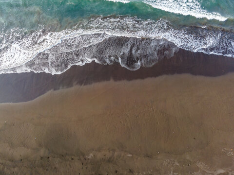 Aerial view of waves crashing onto the dark sand beach in Cilacap, Central Java, Indonesia.