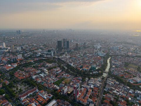 Surabaya, Indonesia - 28 August 2026: Aerial view of the sprawling cityscape featuring dense residential rooftops, modern high-rise buildings, and a winding river under a soft sunset sky.