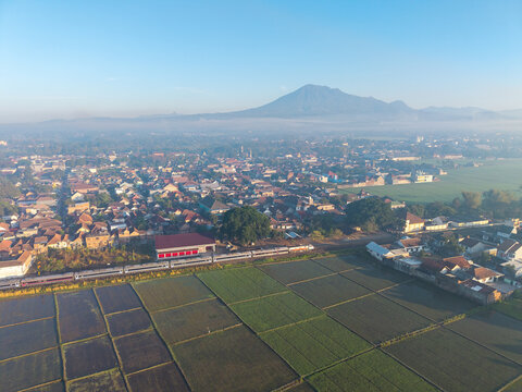 Kepanjen, East Java, Indonesia - 28 August 2026: Aerial view of a train passing through Kepanjen, surrounded by rice fields and buildings, with a mountain in the distance.