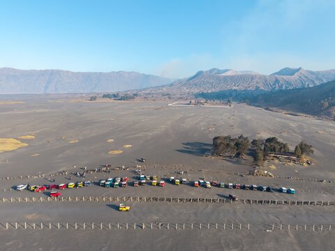 Tosari, Indonesia - 07 August 2026: Aerial view of tourist jeeps parked on the volcanic sand plains of Mount Bromo, with rugged mountain slopes and a clear blue sky in the background.