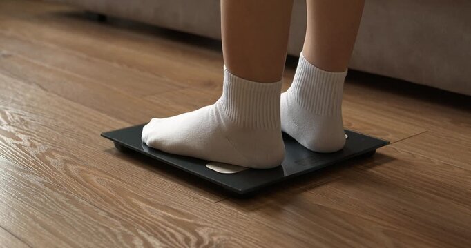Woman feet in white socks stepping on digital electronic scales to check weight on wooden floor.