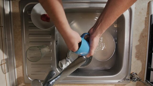 A male doing dishes in a dirty kitchen sink full of water with multiple dishes in natural light.