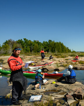 Bearded sea kayaker in red drysuit reading a paper map on granite shoreline, group resting behind, blue sky copy space, Georgian Bay, Ontario