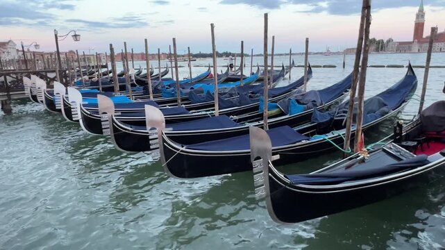 Row of classic Venetian gondolas covered with blue tarps moored at wooden posts on water channel in Venice, Italy.
