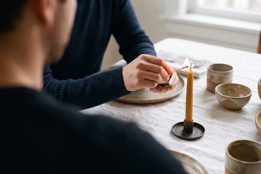 Man lighting a taper candle with a match on a dining table set with ceramic tableware
