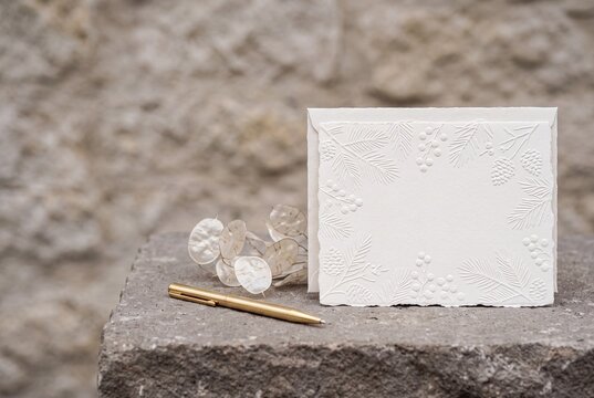 Blank embossed white greeting card mockup with envelope, gold pen, and dried lunaria flowers on rustic stone background