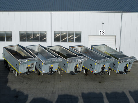Poland - 16 September 2026: Aerial view of a row of five identical grey semi-trailer dump trucks parked in front of a large industrial building with a white wall and a blue roof.
