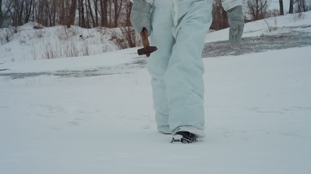 Hazmat researcher trudging across frozen riverbank carrying rusted cross, steady footsteps leave crisp trail, wooded horizon and icy bank, methodical pace and clinical observation, distant outpost