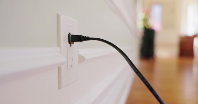 A close up cinematic view of hand plugging black power cord into white electrical outlet on light wall.