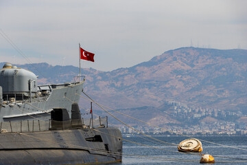 Close-up view of a grey military submarine moored in a harbor with a waving Turkish flag, rusty buoys, and a hazy mountainous coastline with wind turbines in the background. © Selim Aysolmaz