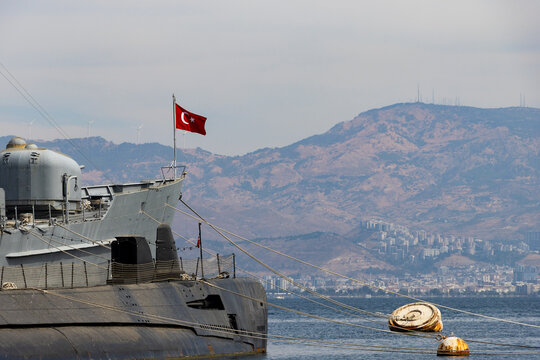 Close-up view of a grey military submarine moored in a harbor with a waving Turkish flag, rusty buoys, and a hazy mountainous coastline with wind turbines in the background.