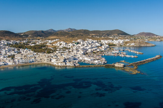 Greece - 21 September 2026: Aerial view of the coastal town of Naoussa on Paros island, featuring white-washed buildings, a small harbor with boats, and clear blue Aegean Sea waters.