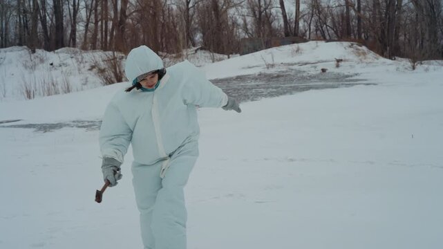 White scientist walking across snowy field, solitary researcher in heavy parka and mittens holding metal hammer near abandoned riverbank, bare trees lining horizon, overcast sky, cold breath visible,