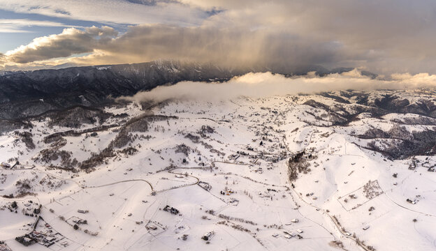 Brasov, Romania - 21 September 2026: Aerial view of the snow-covered landscape of the Carpathian Mountains near Brasov, featuring rolling hills, scattered houses, and dramatic clouds at sunset.