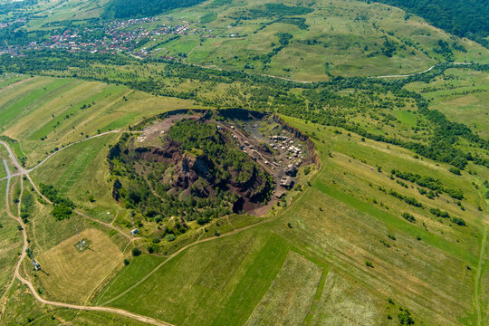 Brasov, Romania - 21 September 2026: Aerial view of the Racos volcanic crater with its unique red and black volcanic rock formations, surrounded by vibrant green meadows and rolling hills.